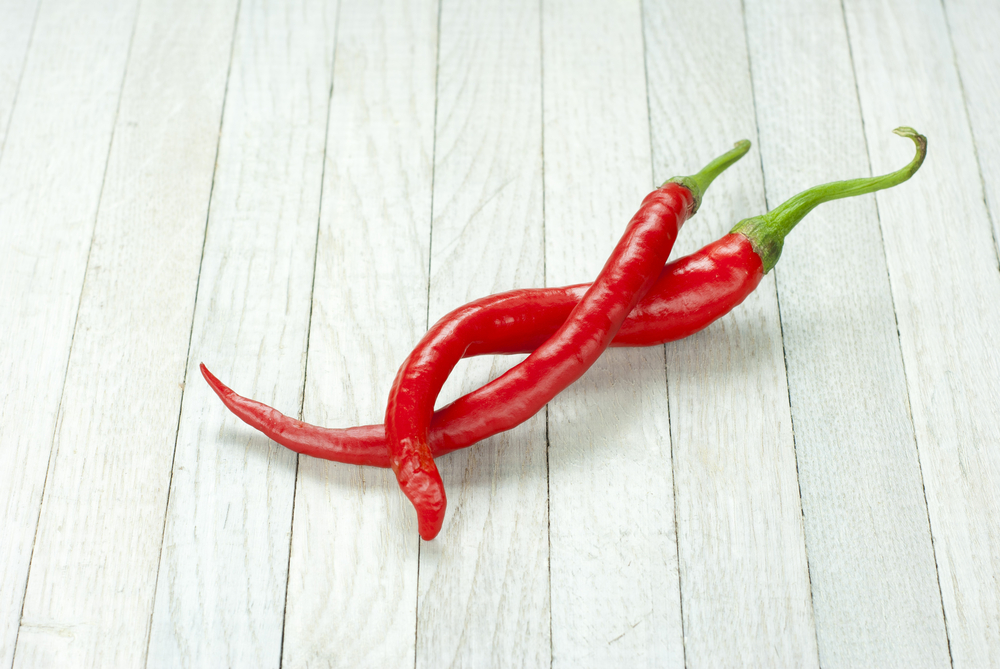 Two red chili peppers, intertwined on a white wooden table.